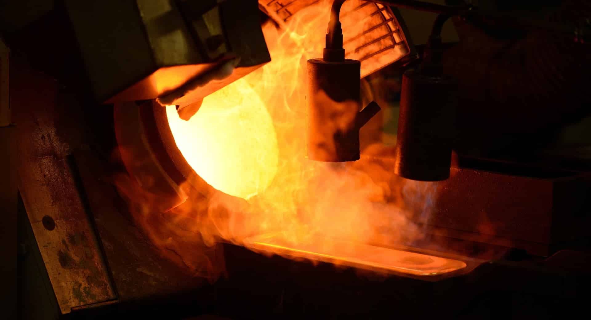 Metal fundido vertido de un crisol a un molde, de color naranja brillante y que produce vapor o humo en una fundición industrial.