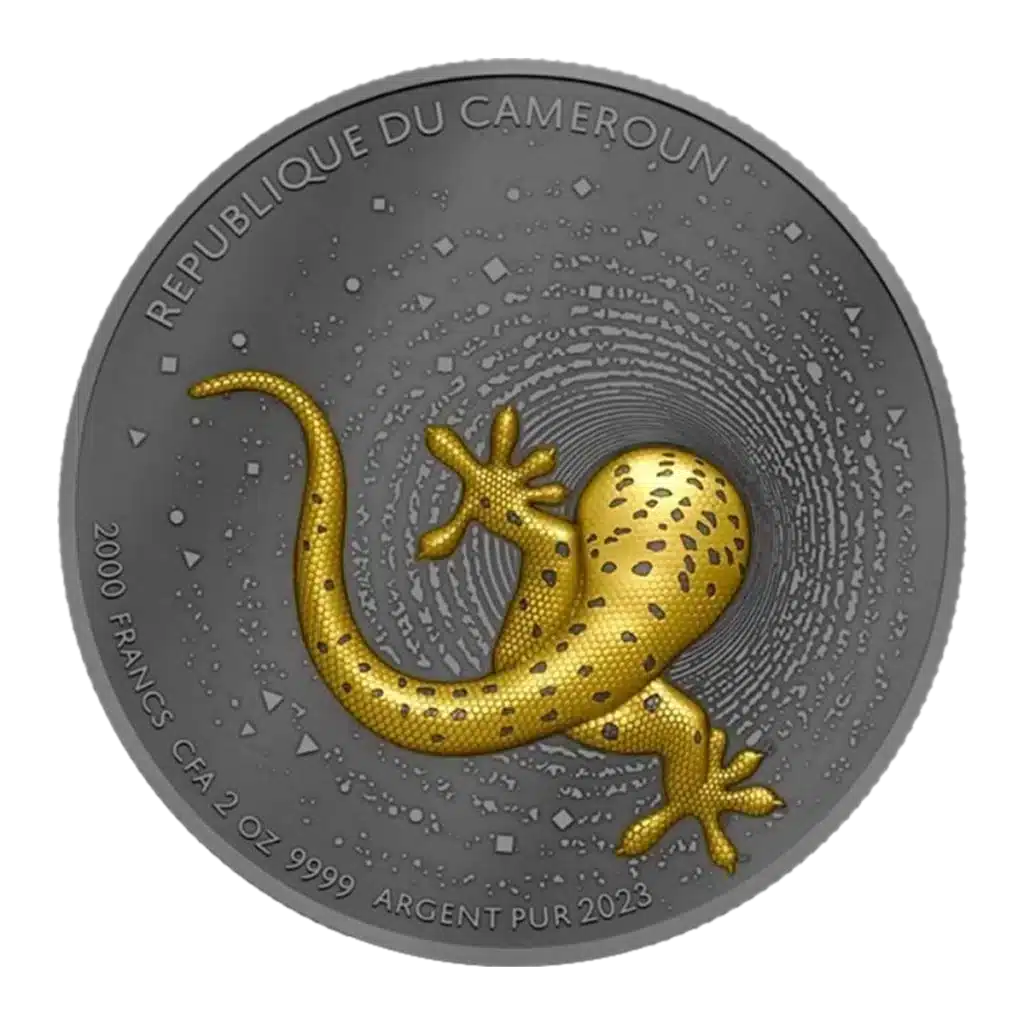 La moneda de plata Camerún 2023 Gecko 2 oz Plata Oscura Dorada UHR muestra un gecko en relieve de color dorado con manchas negras y lleva la inscripción REPUBLIQUE DU CAMEROUN, 2000 FRANCS CFA, 2 OZ .9999 ARGENT PUR 2023.