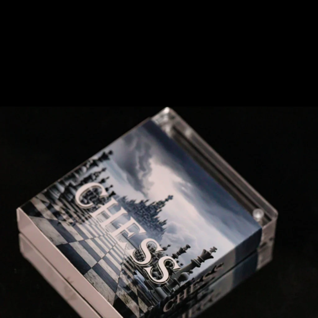 Caja cuadrada transparente con motivo de ajedrez y "Camerún 2024 Ajedrez Internacional 2 oz Plata Acabado Antiguo" en la tapa, sobre una superficie negra. La ilustración de la caja muestra piezas de ajedrez y un tablero de ajedrez.