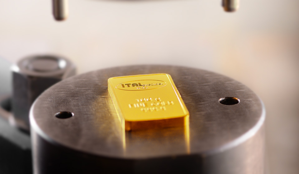 Un lingote de oro con la etiqueta ITAL descansa sobre una plataforma metálica circular, probablemente parte de un proceso de inspección o estampación, con accesorios metálicos visibles en el fondo.