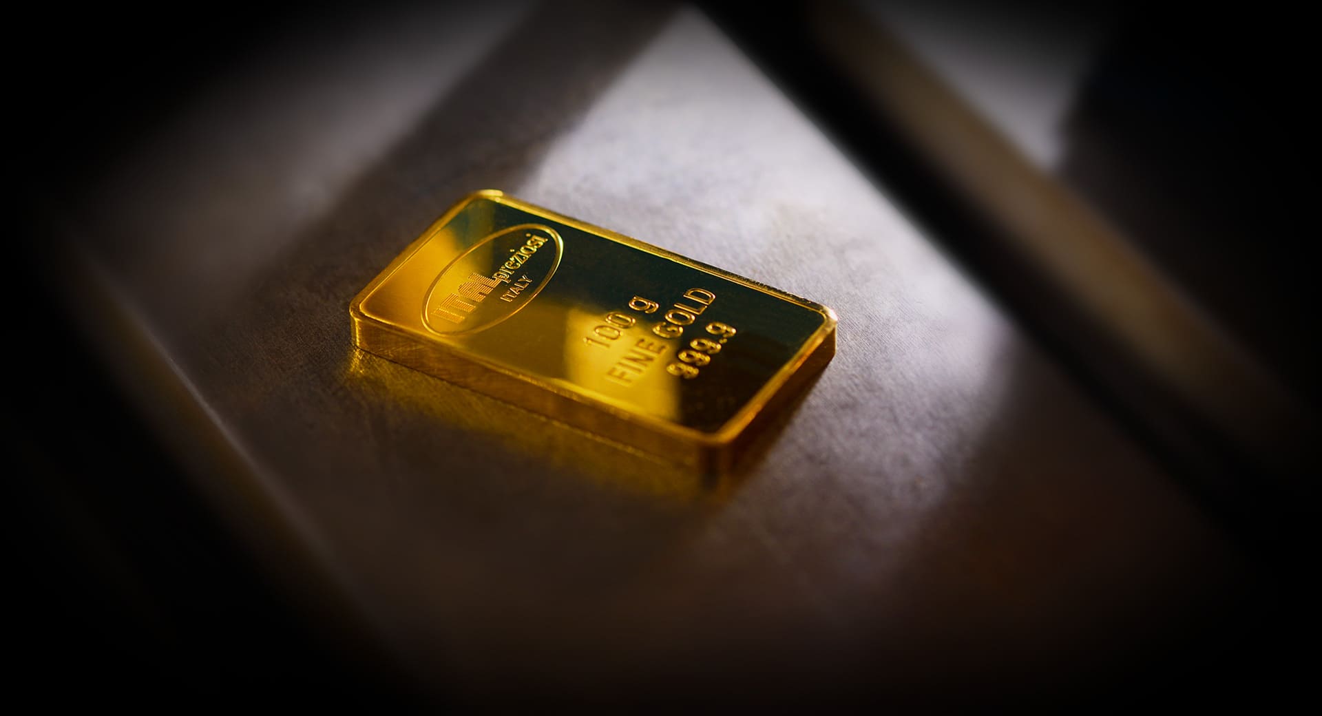 Un único lingote de oro de 100 gramos con marcas grabadas descansa sobre una superficie metálica oscura bajo una iluminación focalizada.