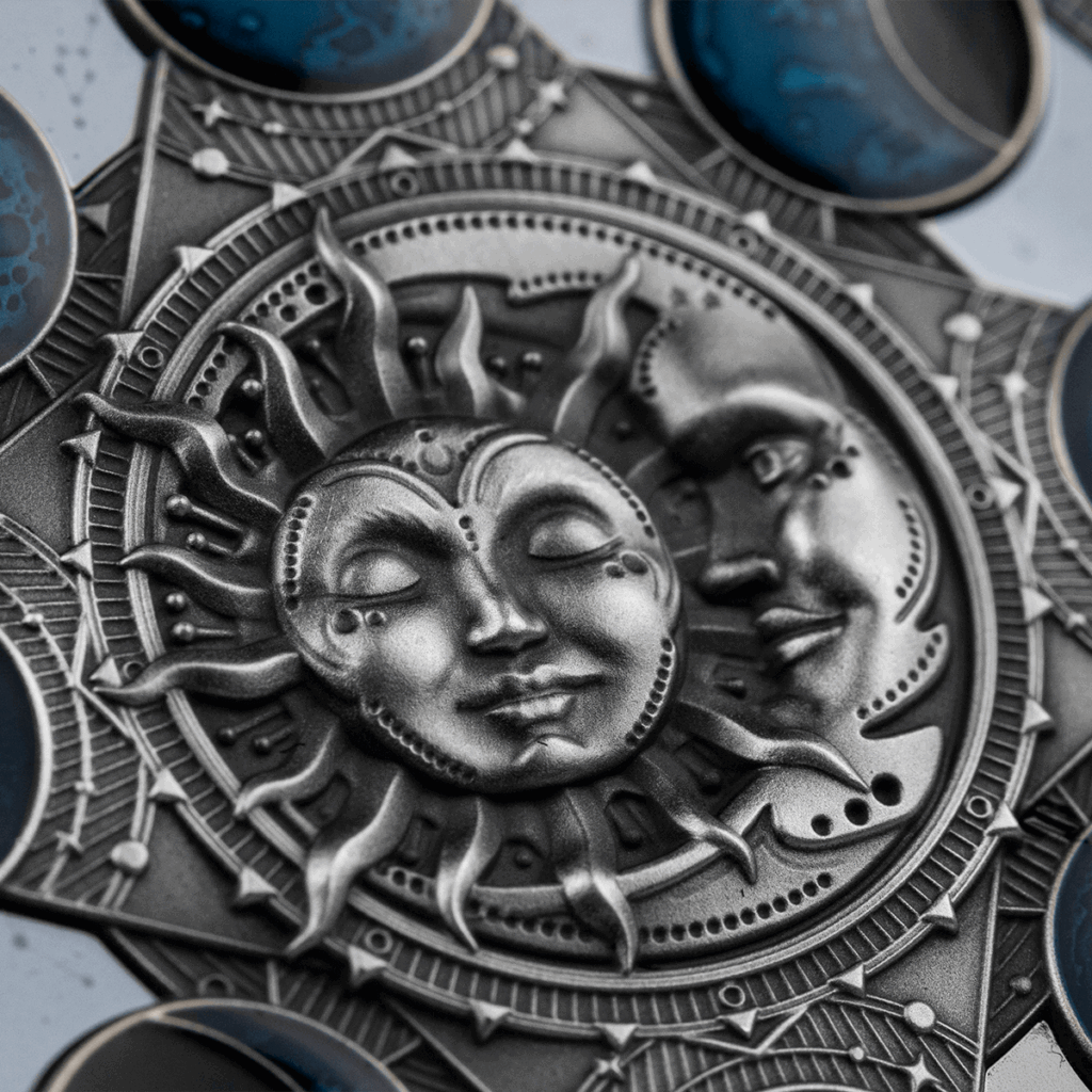 El Calendario Lunar 2 Dólares presenta un relieve metálico con caras estilizadas del sol y de la luna, el sol a la izquierda y la luna a la derecha, ambos rodeados de motivos geométricos y detallados con rasgos serenos y expresivos.