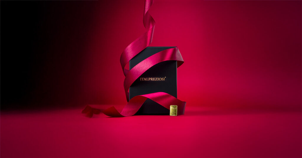 Una caja negra con la inscripción ITALPREZIOSI en letras doradas, que contiene una Barra de Oro, está envuelta con una cinta roja fluida y colocada sobre un fondo rojo intenso. En la base de la caja hay una pequeña etiqueta dorada.