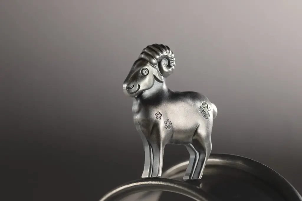 Moneta sagomata Fluffy Silver Sheep 2027 in argento anticato, autoportante, fotografata in piedi su una superficie metallica curva con sfondo grigio sfumato.