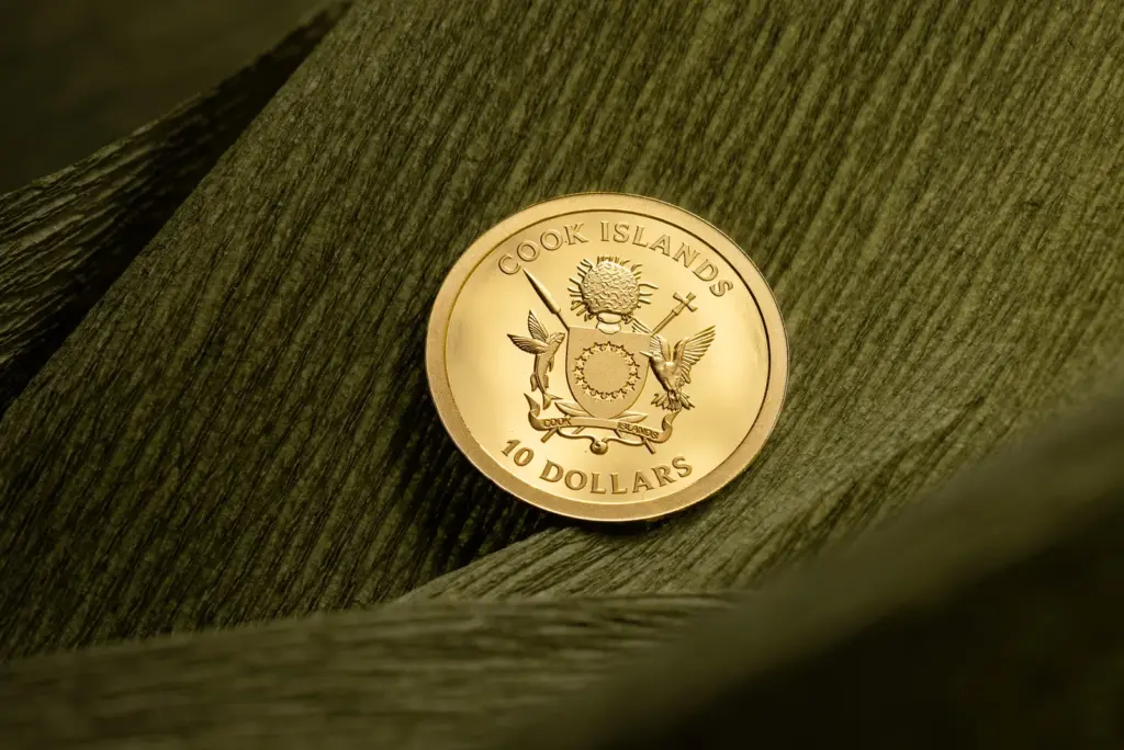 Dritto in oro della moneta Gold Luck 2026 con lo stemma delle Isole Cook, appoggiato su un drappo di carta verde scuro.