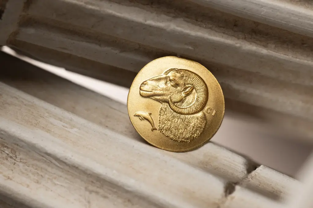 Moneta Krios 0,5 g in oro con la testa di ariete in ultra alto rilievo, appoggiata su una colonna scanalata in pietra chiara di ispirazione classica.