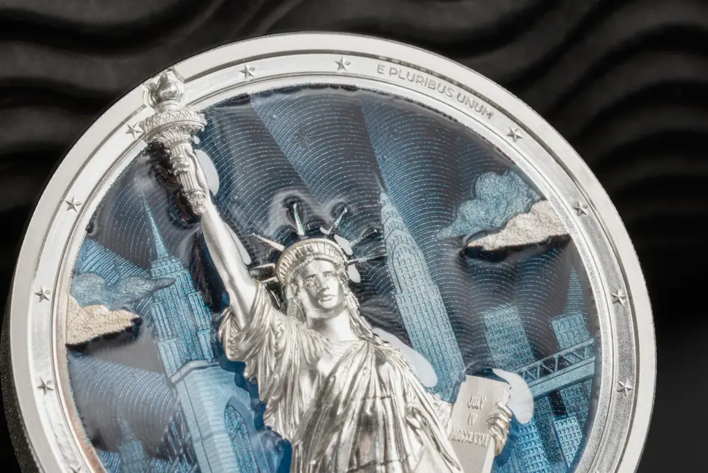 Macro della moneta Timeless Icons - Statua della Liberta 2 oz argento: dettaglio della torcia, della corona e della tavola con la data JULY IV MDCCLXXVI, sullo sfondo blu smaltato inciso a guilloche.